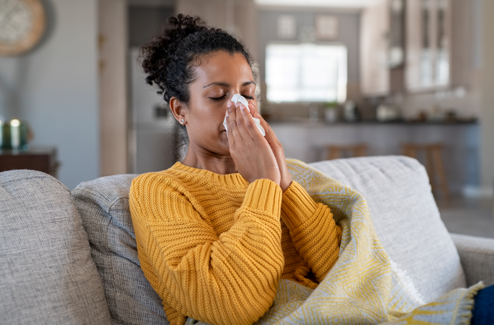 A woman sits on a couch wrapped in a blanket and holds a tissue up to her nose